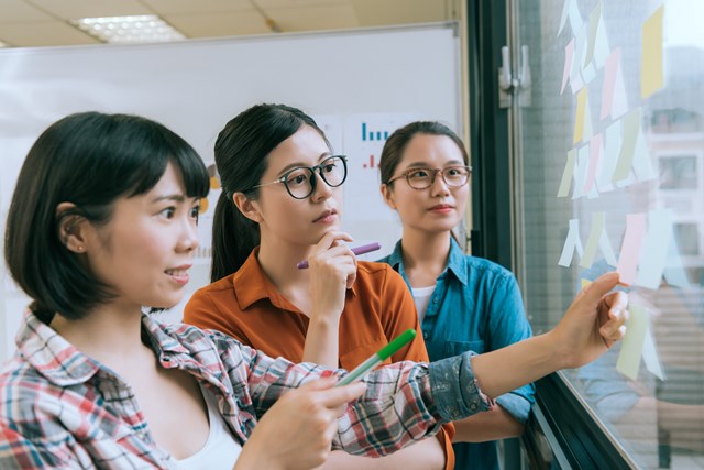 Professional young female designer team meeting on glass wall and looking at many sticky memo paper thinking discussing design case planning.